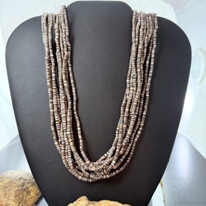 May include: A multi-strand necklace composed of small, light brown and white beads. The necklace is displayed on a black mannequin bust. The beads are strung together to create a layered effect, suitable for casual or formal wear.