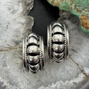 May include: A pair of silver hoop earrings. Each earring features a series of rounded, polished silver beads separated by open spaces. The edges are detailed with a twisted rope design, creating a textured border. The earrings are displayed on a light-colored, patterned surface.