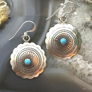 Könnte beinhalten: Ein Paar silberfarbene Ohrringe mit einem türkisfarbenen Mittelstein. Die Ohrringe haben einen gewellten Rand und ein detailliertes Blumenmuster. Die Ohrhaken sind silberfarben und gebogen.