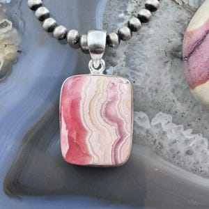 May include: A rectangular pendant featuring a banded pink and cream stone set in a silver-toned metal frame, suspended from a necklace composed of round, polished silver-toned beads that create a continuous chain around the central stone piece.
