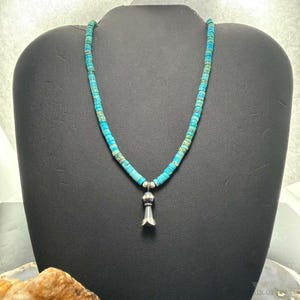 May include: A necklace featuring a strand of small, cylindrical turquoise beads in shades of blue and green. The necklace includes a central silver-toned metal pendant shaped like a small, smooth bell or cone, suspended from a silver-toned bead.