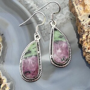 May include: A pair of teardrop-shaped earrings featuring polished zoisite stones with distinct patches of vibrant magenta and mossy green. Each stone is encased in a twisted silver-tone metal border and hangs from a simple silver-tone hook wire.