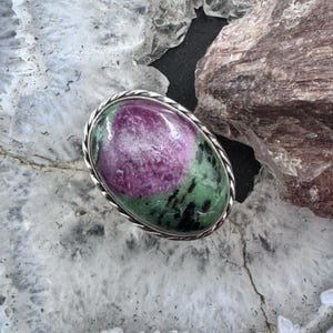 May include: An oval ring featuring a polished ruby zoisite gemstone with a prominent magenta center surrounded by green and black mineral inclusions, set within a twisted silver-toned metal frame that follows the oval shape of the stone.