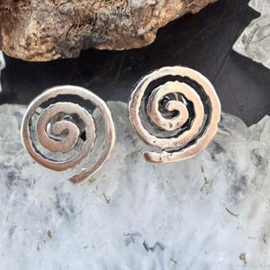 May include: A pair of silver-toned spiral stud earrings. Each earring features a swirling design with a raised, textured edge. The earrings are displayed on a light-colored, textured surface.