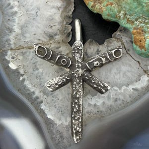 May include: A dark gray, textured metal pendant shaped like a stylized dragonfly with a vertical body and two horizontal wings. The wings feature circular indentations, and the entire surface has a rough, pitted finish with a silver-toned bail.