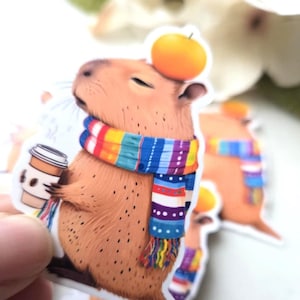 May include: A brown capybara wearing a colorful striped scarf and holding a cup of coffee. The capybara has an orange fruit on its head.