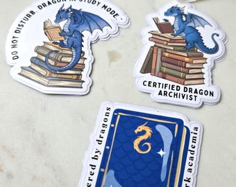 May include: Three stickers featuring dragons and books. One sticker reads "Do Not Disturb. Dragon in Study Mode." Another says "Certified Dragon Archivist." The third sticker shows a book with a dragon and the text "Powered by dragons and dark academia."