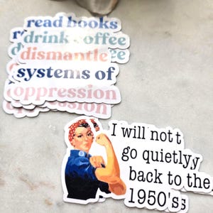 May include: A set of stickers with feminist slogans, including "read books", "r'drink offee", "dismantle systems of oppression", and "I will not go quietly back to the 1950's". The sticker with the slogan "I will not go quietly back to the 1950's" features a vintage illustration of Rosie the Riveter.