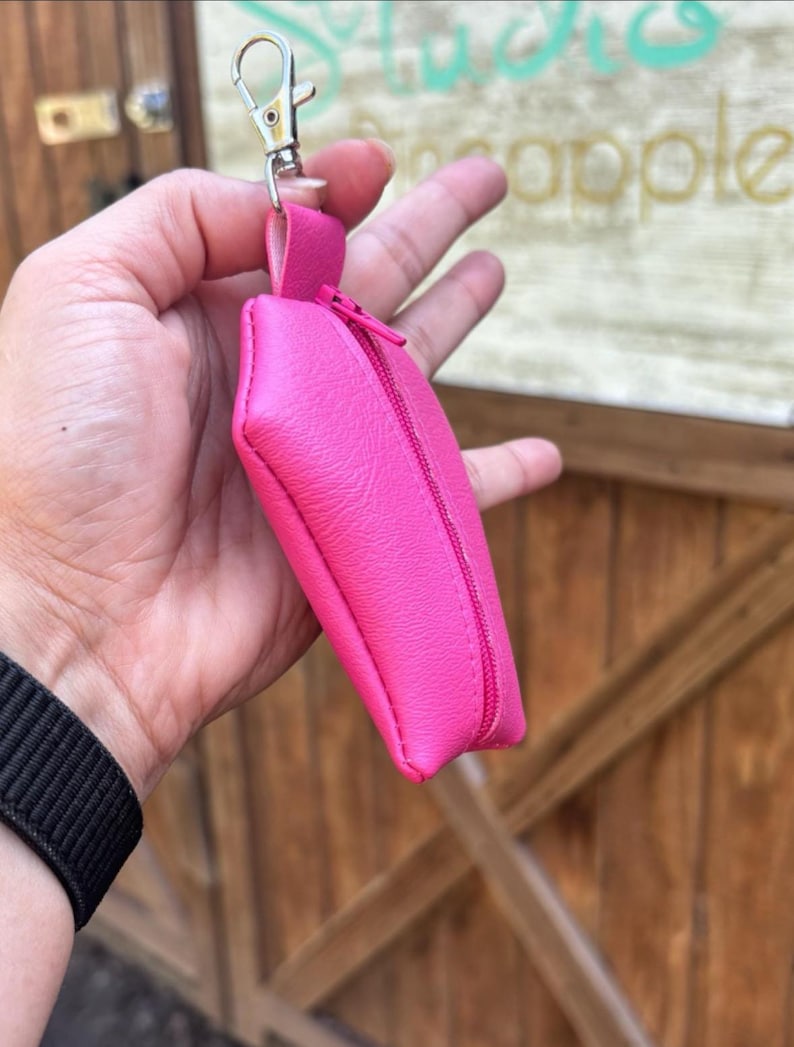 Pode incluir: Uma bolsa rosa brilhante com z&iacute;per e um chaveiro prateado. A bolsa &eacute; feita de um material macio e texturizado.