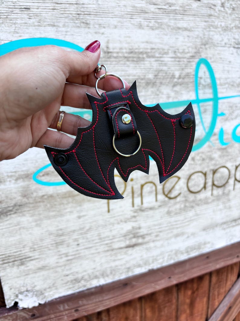 May include: A black leather bat-shaped keychain with red stitching and a silver ring. The keychain has a small black button on the back.