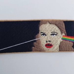 May include: Embroidered patch featuring a woman's face with a rainbow prism behind her ear. The background is black and the patch has a beige border.