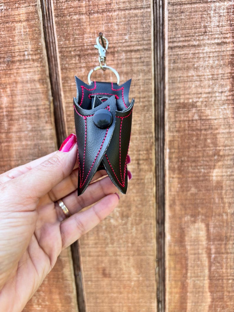 May include: A black leather keychain with a red stitch detail and a black button closure. The keychain is shaped like a small pouch.