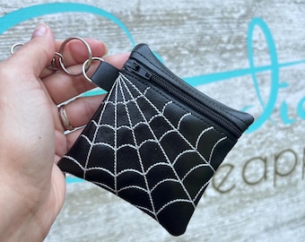 May include: Black coin purse with a white spiderweb design, featuring a zipper closure and a key ring. The small, square-shaped pouch is held by a hand against a blurred background.