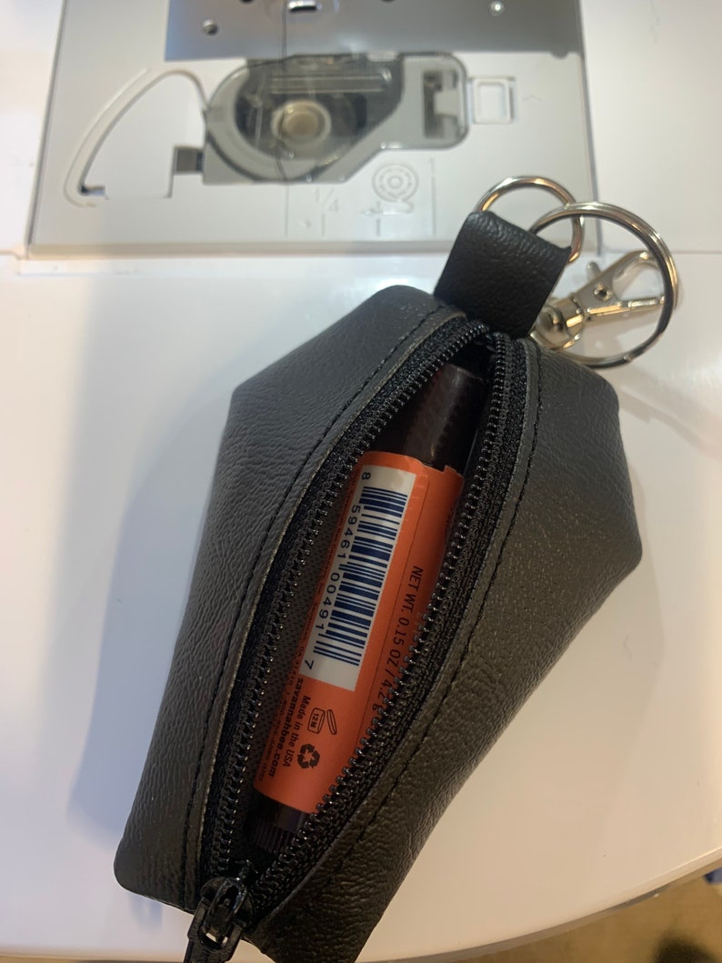 Pode incluir: Uma bolsa de couro preto com z&iacute;per e um chaveiro anexado. A bolsa est&aacute; aberta e cont&eacute;m um pequeno tubo laranja de b&aacute;lsamo labial com o texto "NET WT. 0.15 OZ/4.2 g" e um c&oacute;digo de barras. O b&aacute;lsamo labial &eacute; feito nos Estados Unidos.