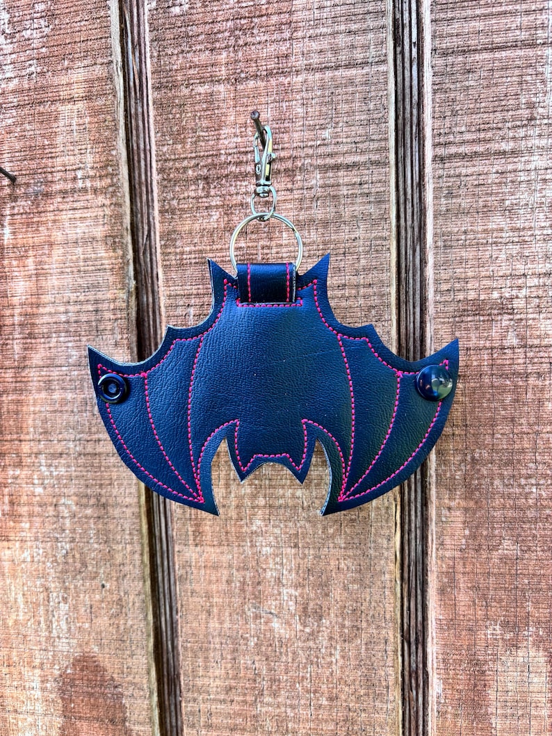 May include: A black leather bat-shaped keychain with pink stitching and a silver metal clasp. The keychain is hanging from a nail on a wooden surface.