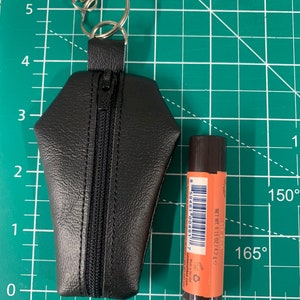 Pode incluir: Um chaveiro de couro preto em forma de caix&atilde;o com fecho de correr e um aro de chave prateado. Um pequeno tubo de b&aacute;lsamo labial laranja com c&oacute;digo de barras e o texto "NET WT. 0.15 OZ/4.2 g" est&aacute; ao lado do chaveiro.