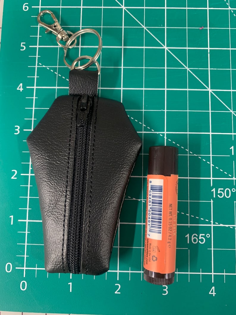 Pode incluir: Um chaveiro de couro preto em forma de caix&atilde;o com fecho de correr e um aro de chave prateado. Um pequeno tubo de b&aacute;lsamo labial laranja com c&oacute;digo de barras e o texto "NET WT. 0.15 OZ/4.2 g" est&aacute; ao lado do chaveiro.