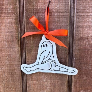 May include: A white ghost-shaped ornament with black details and an orange ribbon. The ghost is in a split position. The ornament is hanging against a wooden background. Halloween-themed decor.