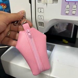 Pode incluir: Uma bolsa rosa com z&iacute;per em forma de caix&atilde;o. A bolsa est&aacute; pendurada em um chaveiro prateado.