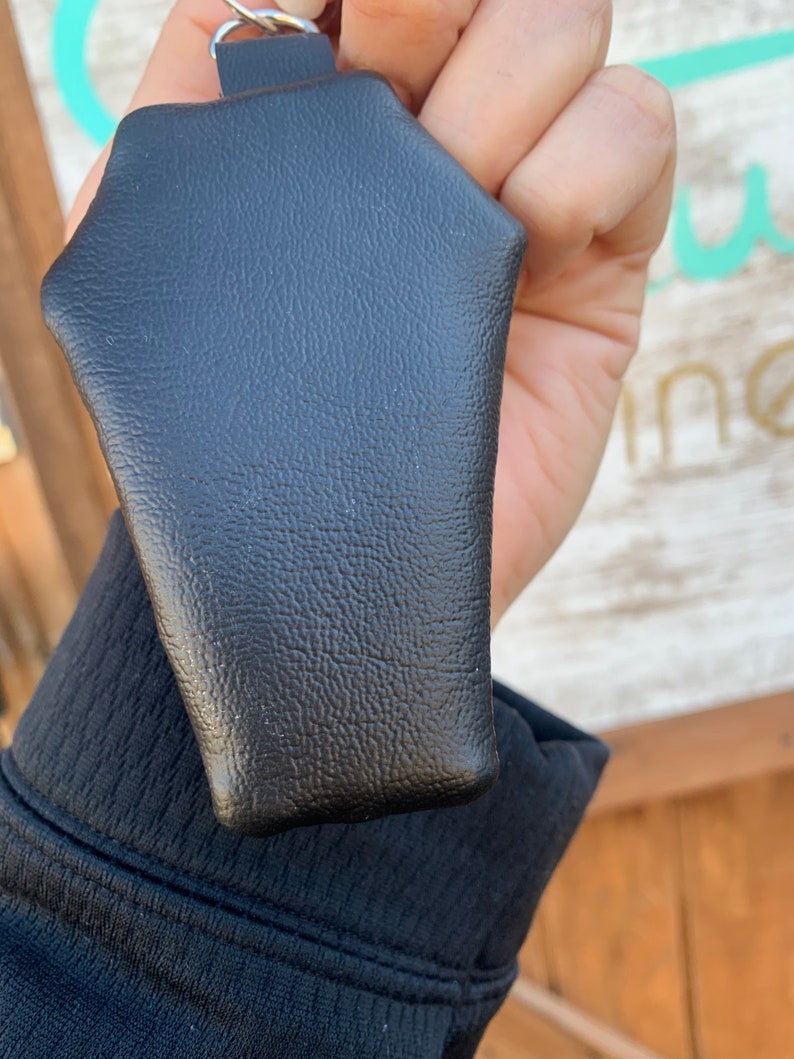 Pode incluir: Um chaveiro de couro preto em forma de caix&atilde;o com um anel prateado.