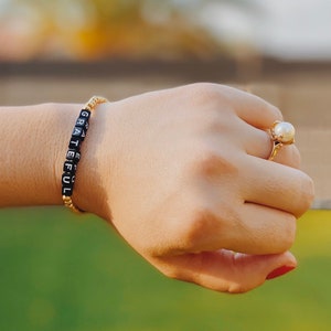May include: A gold bracelet with black beads spelling out the word "grateful". The bracelet is worn on a person's wrist, with a gold ring on the same hand.