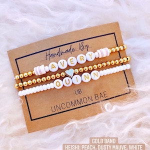 May include: Three gold bead bracelets with white letter beads spelling out the names Avery and Quinn. The bracelets are on a brown card with the text "Handmade By", "UB", "Uncommon Bae", "Gold Band", and "Heishi: Peach, Dusty Mauve, White".