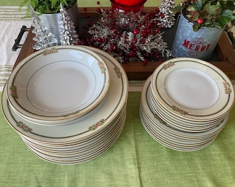 May include: A stack of 12 white ceramic dinner plates with a gold rim and a floral design. The plates are stacked on a green tablecloth.
