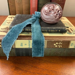 May include: A stack of antique books with teal ribbon and a decorative glass sphere. The books are bound in brown and green leather, with gold lettering and floral designs. The glass sphere has a swirling pattern of white and purple.