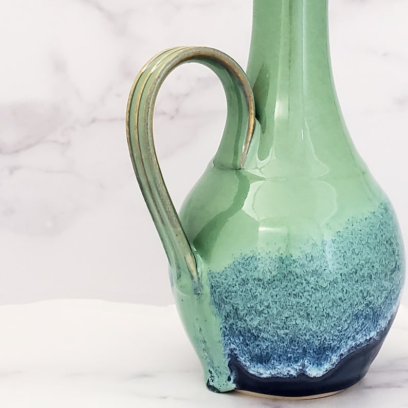 May include: A green ceramic pitcher with a blue and white glaze. The pitcher has a handle with gold accents.