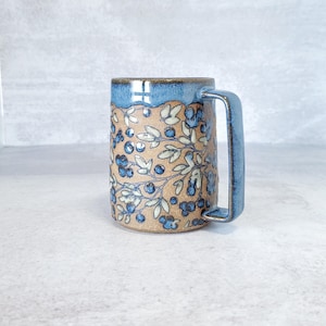 Peut inclure: Une tasse en céramique faite à la main avec une anse et un bord bleus. La tasse présente un corps beige avec un motif détaillé de baies et de feuilles bleues. La tasse mesure environ 13 cm de haut.