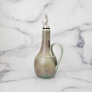 May include: A green ceramic oil and vinegar bottle with a silver pour spout and a handle. The bottle has a mottled glaze and a rustic look.