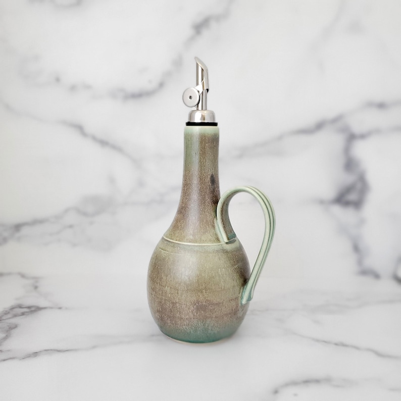 May include: A green ceramic oil and vinegar bottle with a silver pour spout and a handle. The bottle has a mottled glaze and a rustic look.
