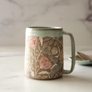 May include: A ceramic mug with a light green glaze and a floral design. The design features pink and white flowers and leaves on a brown background.