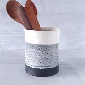 May include: A ceramic kitchen utensil holder with a white top, a textured gray middle section, and a dark gray base. Three wooden spoons are inside the holder. The holder is cylindrical and stands upright.