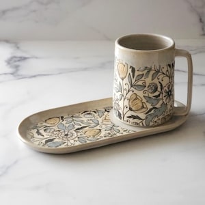 May include: A ceramic mug and matching tray with a floral pattern in shades of gray, blue, and yellow. The mug has a handle and the tray is oval shaped.