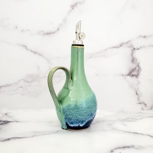 May include: A green and blue ceramic oil and vinegar bottle with a stainless steel pour spout and a handle.