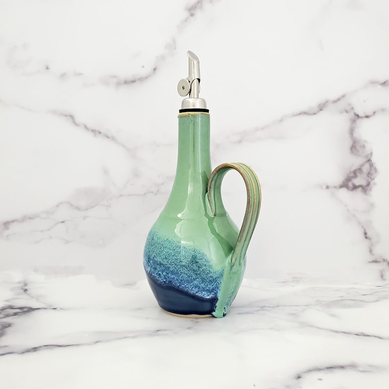 May include: A green and blue ceramic oil and vinegar bottle with a stainless steel pour spout and a handle.