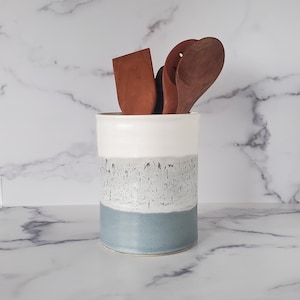 May include: A white ceramic utensil holder with a grey and blue glaze. The holder is filled with wooden kitchen utensils.