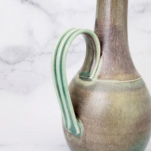 May include: A ceramic pitcher with a green and brown glaze. The pitcher has a wide, round body and a narrow neck. The handle is green and has a striped design.
