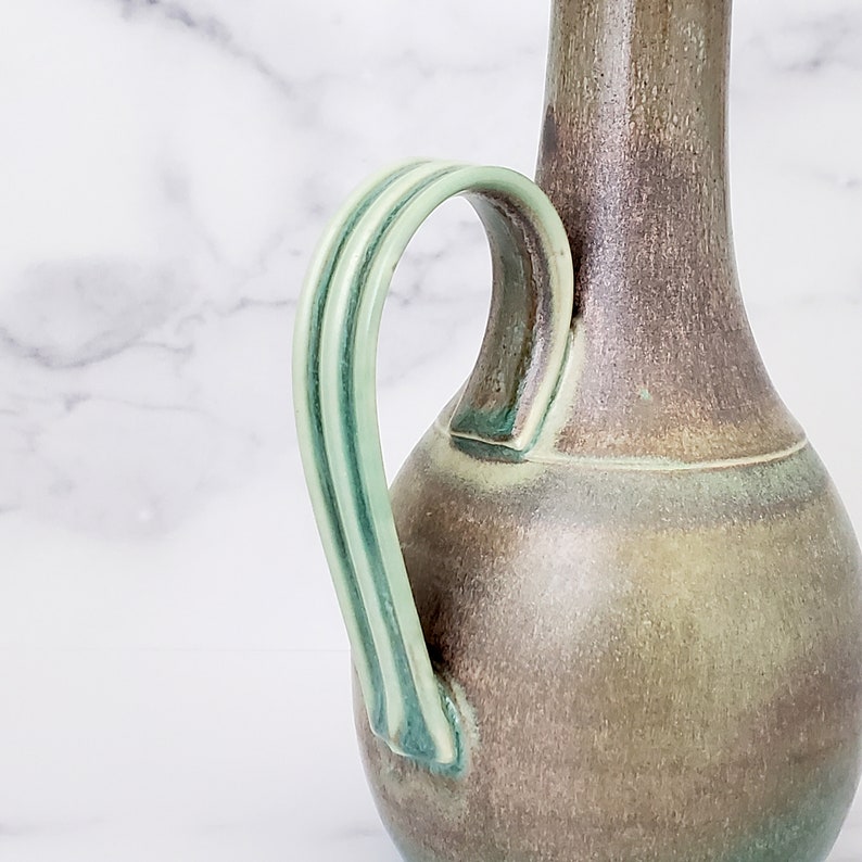 May include: A ceramic pitcher with a green and brown glaze. The pitcher has a wide, round body and a narrow neck. The handle is green and has a striped design.