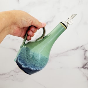 May include: A green and blue ceramic oil and vinegar bottle with a stainless steel pour spout and a handle.