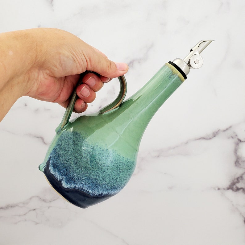 May include: A green and blue ceramic oil and vinegar bottle with a stainless steel pour spout and a handle.
