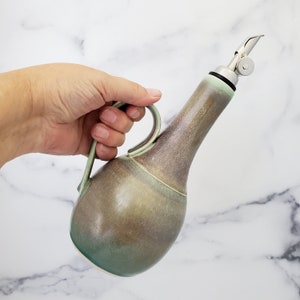 May include: A ceramic olive oil bottle with a green and brown glaze. The bottle has a pour spout with a stainless steel lid and a handle.