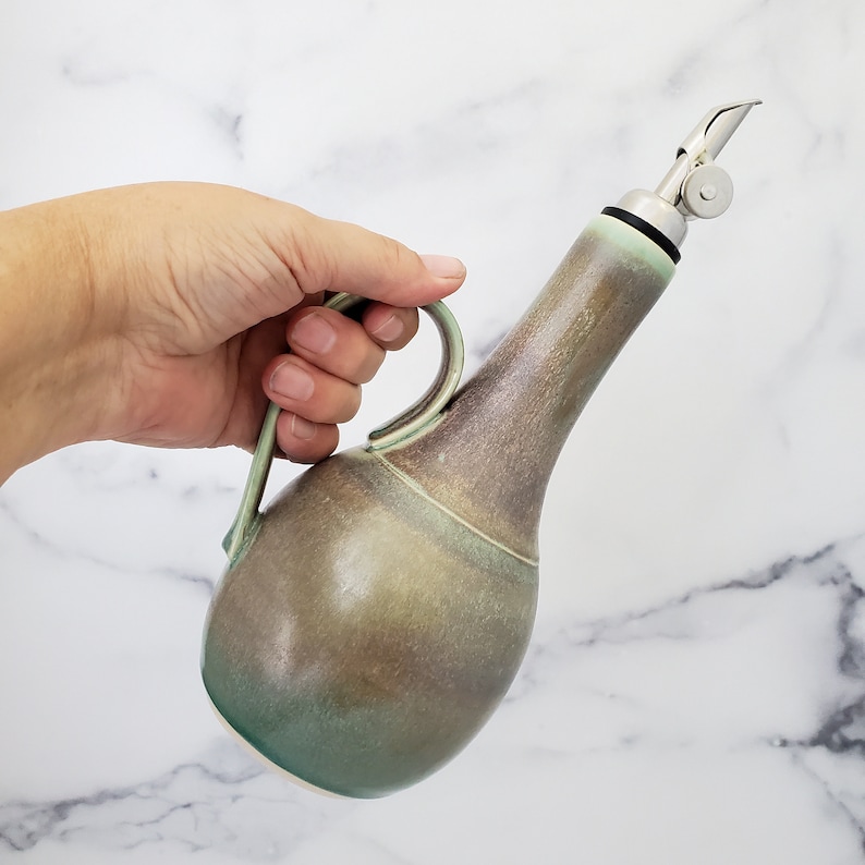 May include: A ceramic olive oil bottle with a green and brown glaze. The bottle has a pour spout with a stainless steel lid and a handle.