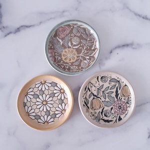 May include: Three ceramic trinket dishes with floral patterns. The dishes are round and have a white background with different coloured floral designs. The first dish has a light blue background with a brown floral pattern. The second dish has a light yellow background with a white floral pattern. The third dish has a light pink background with a black floral pattern.