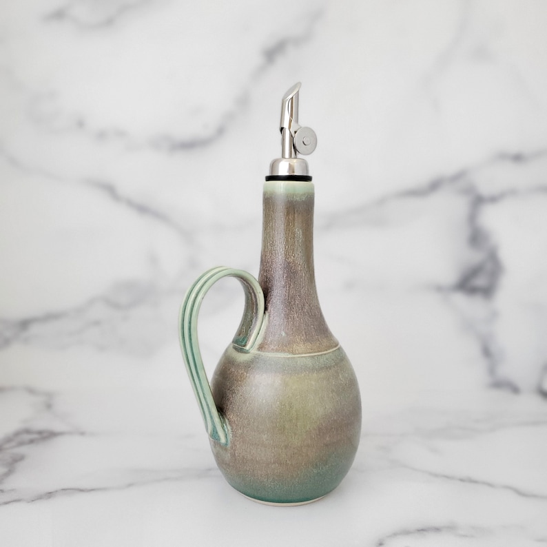 May include: A ceramic olive oil bottle with a green and brown glaze. The bottle has a spout and a handle. It is sitting on a white marble surface.