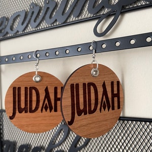 May include: A pair of wooden earrings with the name "JUDAH" engraved on each earring. The earrings are hanging on a black metal earring holder.