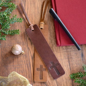 May include: Two brown leather bookmarks with a cross design. The bookmarks are on a wooden surface with a red notebook and a black pen.