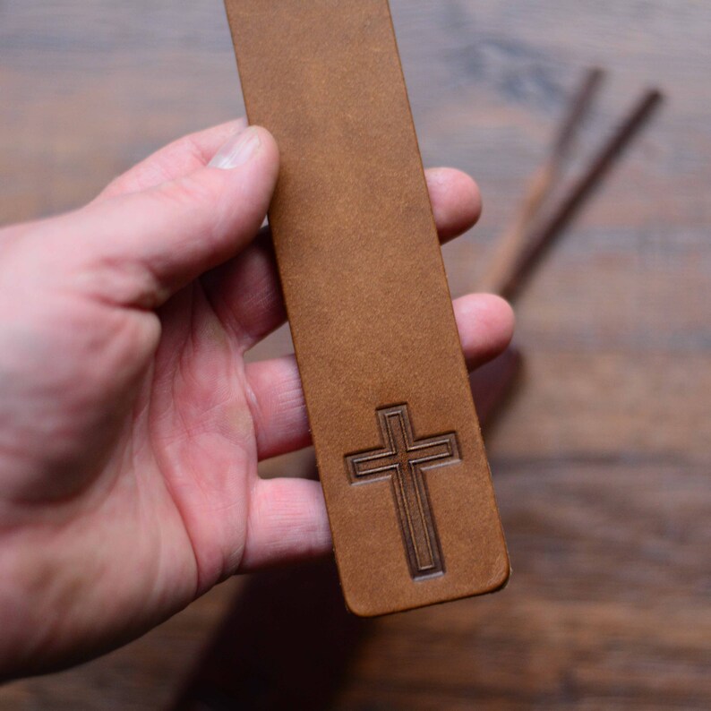 May include: A brown leather bookmark with a cross design. The bookmark is held in a hand.
