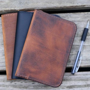 May include: Three rectangular leather notebook covers in tan, dark brown, and black with visible grain textures and rounded corners, accompanied by a clear plastic ballpoint pen with a black grip and a black clip positioned to the right side.
